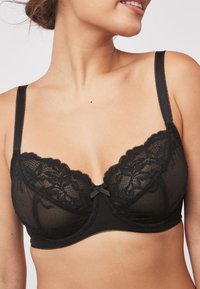 Black lace bra with sheer mesh cups, featuring floral patterns, a scalloped edge, and a small bow accent at the center. Adjustable straps.