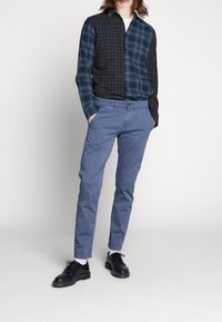 Károvaná košile v tmavě modré a zelené barvě, zapínání na knoflíky; kombinovaná se světle modrými chino kalhotami a černými botami. Minimalistický vzhled s čistými liniemi.