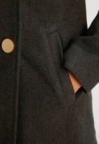 Mørkegrå uldfrakke med en glat overflade, prydet med en gylden rund knap og sidelommer, der understreger det klassiske design.