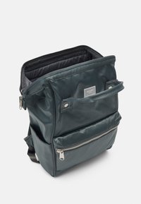 Ciemnozielony plecak z nylonu z zamykaną na zamek główną komorą, przednią kieszenią i srebrnymi dodatkami. Gładka tekstura, prostokątny kształt.