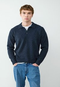Granatowy, długi sweter polo z teksturowanym splotem, z trzema guzikami i ściągaczami na rękawach, noszony na białej koszulce i niebieskich dżinsach.
