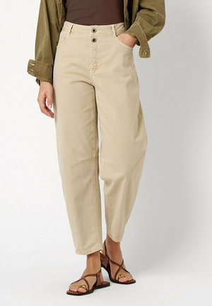Person wearing beige high-waist pants with buttons, brown top, olive green jacket, and brown open-toe heeled sandals standing with one hand in pocket.