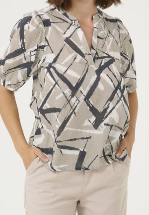 Vrouw in een beige, zwart en witte gestreepte V-hals kortekmouwen blouse met handen in de lichte beige broekzakken.