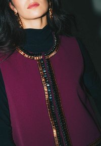 Top sans manches de couleur bordeaux avec un col orné de détails décoratifs noirs et dorés brillants. Texture de tissu lisse avec une coupe structurée.
