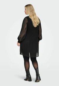 Černé plisované šaty s průhlednými dlouhými rukávy, doplněné černými punčochami a kotníkovými botami na podpatku. Hladká textura a volný střih.