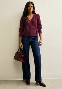 Maglione avvolgente bordeaux con maniche lunghe, jeans svasati blu scuro e una piccola borsa a tracolla bordeaux. Modella che indossa stivaletti con tacco a punta.