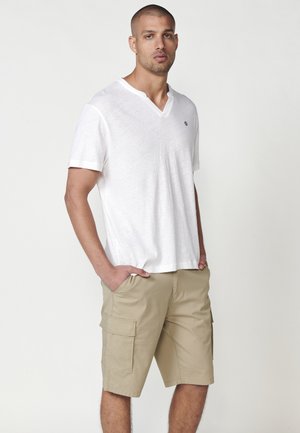 Camiseta blanca de manga corta y cuello en V con una textura sutil y un pequeño emblema en el pecho, combinada con pantalones cortos tipo cargo beige que tienen bolsillos laterales.
