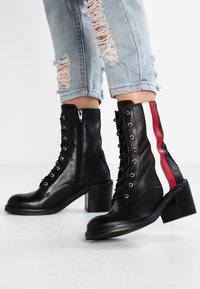 Bottines en cuir noir à lacets et fermeture éclair sur le côté, ornées de rayures rouges et blanches, portées avec un jean déchiré bleu clair.