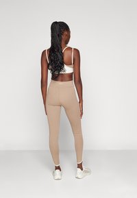 Beige leggings med en tät passform och hög midja. I kombination med en ljusbrun sport-bra. Modellen har långt, flätat hår. Vita sneakers.