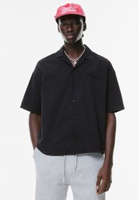 Cămașă neagră cu mânecă scurtă și nasturi, cu un croi lejer și o textură subtilă. Pur tată cu pantaloni scurți gri și o șapcă roz cu logo alb.