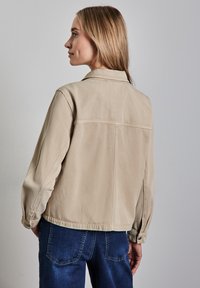 Veste en denim beige clair avec un col classique, des poignets boutonnés et un détail de découpe au dos. Associée à un jean bleu, montrant une coupe décontractée.