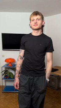 Mladić s tetovažama na obje ruke nosi crnu majicu i tamne hlače, stoji u sobi s policom, lampom i televizorom na zidu.