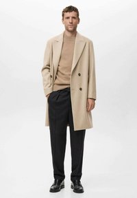 Abrigo beige de doble botonadura sobre un suéter de punto color canela, combinado con pantalones negros plisados y zapatos negros, frente a un fondo sencillo.