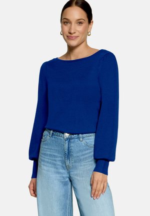 Femme aux cheveux foncés portant un haut bleu à manches longues et un jean bleu clair, debout devant un fond uni clair, souriant légèrement.