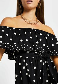 Kvinna i svart off-shoulder klänning med prickar och volangkrage, guldhalsband och stora hoopörhängen.