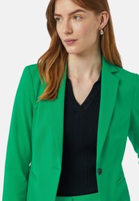 Grüner, taillierter Blazer mit glatter Textur, einem Ein-Knopf-Verschluss und Reverskragen, kombiniert mit einem schwarzen, gerippten V-Ausschnitt-Oberteil.