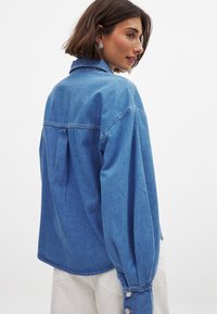 Hellblaue Jeanshemd mit langen Ärmeln, einer Rückenfalten und geknöpften Manschetten. Hat einen kragenähnlichen Ausschnitt und einen lässigen Schnitt.