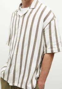 Camisa de manga corta en blanco con rayas verticales marrones, con detalles sutiles en los bordes y un corte relajado. Tejido texturizado.