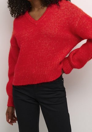 Kvinde iført en klart rød, blød V-udskærings sweater med lange ærmer og sorte bukser, med den ene hånd hvilende på hoften.