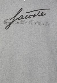 Sudadera gris con un bolsillo canguro en el frente, un gran logo negro de "Lacoste" y cinco diseños de cocodrilos contorneados arriba. Textura de tela suave.