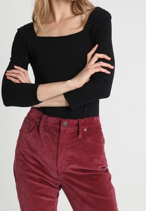 Mujer con los brazos cruzados que lleva una camiseta negra de manga larga con escote cuadrado y pantalones altos de pana rojos.