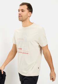 Lys beige t-shirt, rund hals, korte ærmer, med topografisk mønster og "camel active" tekst i rød. Blød bomulds tekstur.