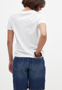 Camiseta de algodón blanca con mangas cortas, combinada con pantalones cortos de mezclilla azul oscuro. Los pantalones cortos tienen dos bolsillos traseros y un lavado ligeramente desvanecido.