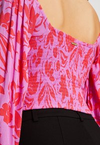 Haut à motif floral en rose et rouge, présentant un design froncé, des manches hors-épaule, et un petit logo doré en accent.