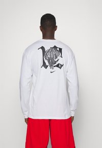 Weißes Langarm-T-Shirt mit einem großen schwarzen Grafikdesign auf dem Rücken und einem Nike-Logo in der Nähe des Saums. Getragen mit roten Shorts.