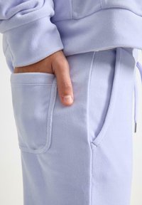 Lys lilla joggingbukser i fleece med sidelomme og justerbar talje med snøre. Stoffet er blødt med en glat tekstur og synlige syningsdetaljer.