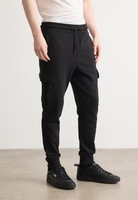 Schwarze Jogginghose aus weichem Stoff, mit Gummizug, Seitentaschen und einer Cargo-Tasche mit Druckknopfverschluss. Kombiniert mit schwarzen Sneakers.