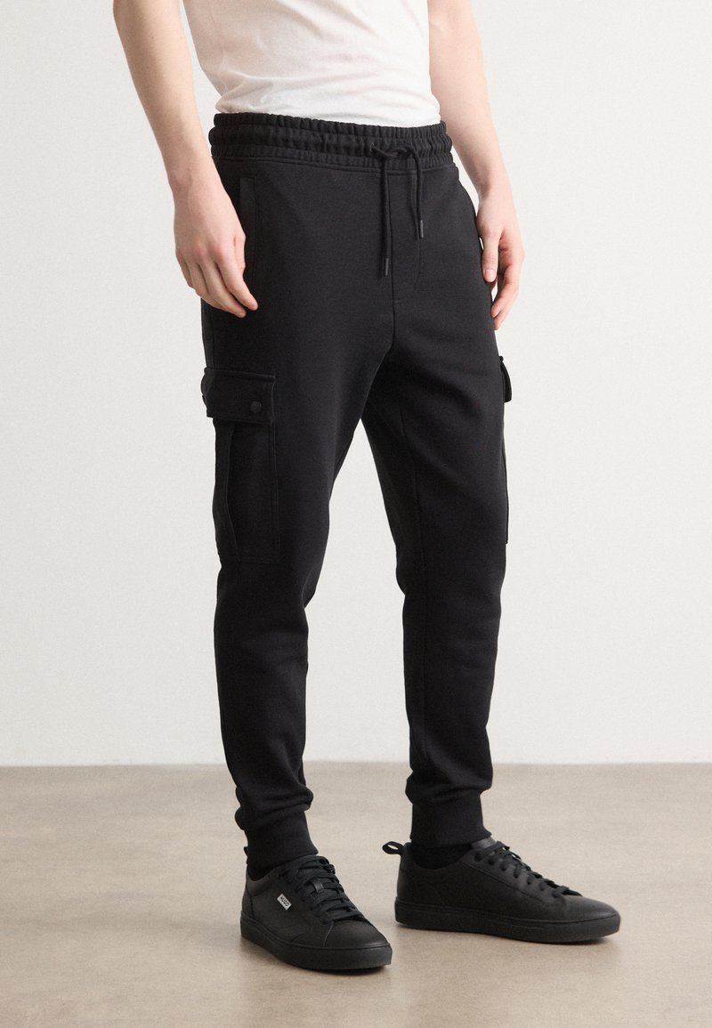 Schwarze Jogginghose aus weichem Stoff, mit Gummizug, Seitentaschen und einer Cargo-Tasche mit Druckknopfverschluss. Kombiniert mit schwarzen Sneakers.