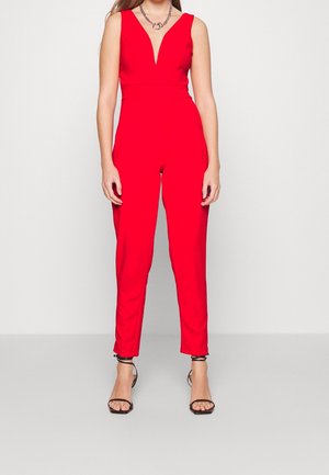 Kvinde iført en ærmeløs rød jumpsuit med dyb V-udskæring og sorte åbne hæle, stående mod en ensfarvet lys baggrund.