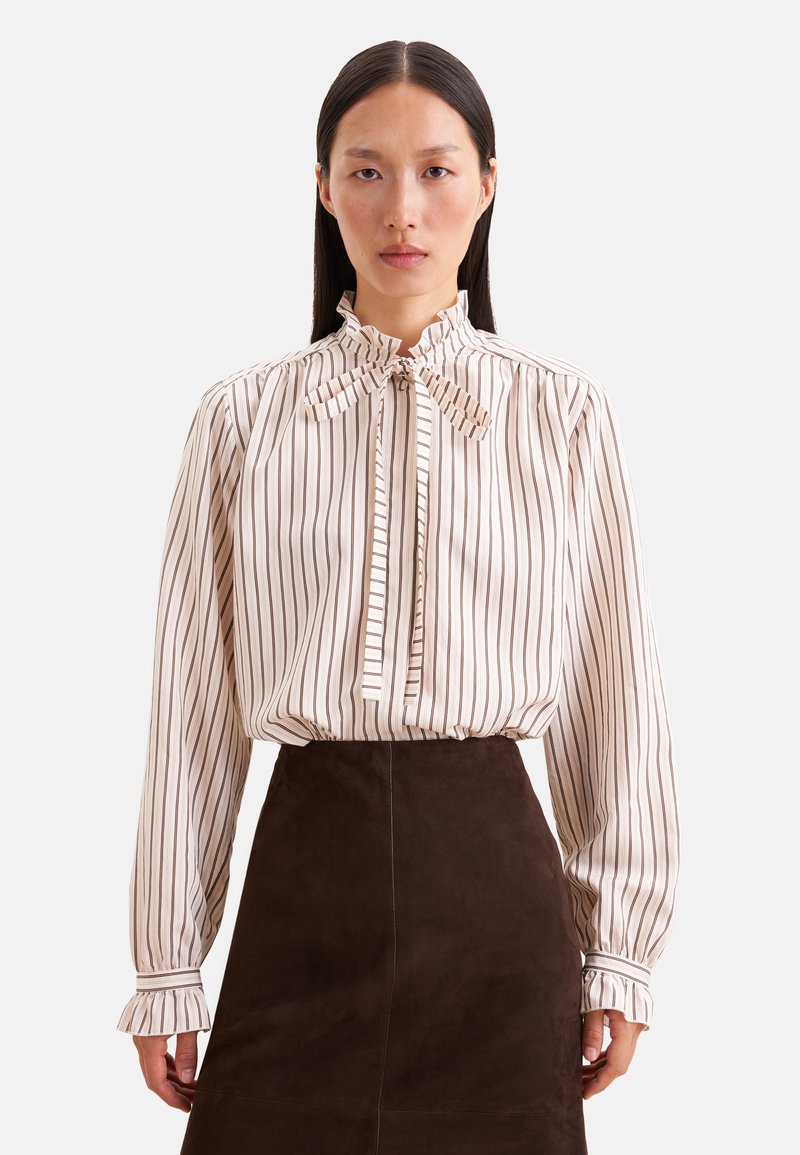 Femme portant une blouse blanche à rayures verticales marron et un col noué, associée à une jupe marron foncé taille haute.