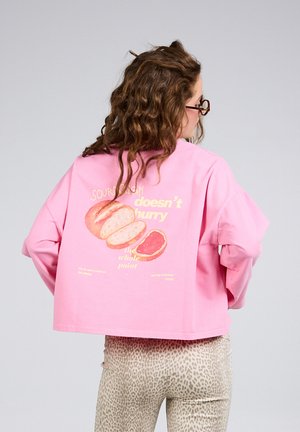 Femme aux cheveux bouclés portant un haut rose à manches longues avec une illustration de pain et du texte, associée à un pantalon léger imprimé léopard, vue de dos.