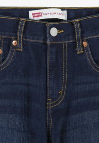 Vaqueros de mezclilla azul oscuro con ajuste slim, botones de cobre, costuras amarillas y una etiqueta que dice "Levi's 512 Slim Tape".