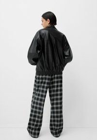 Jachetă bomber din piele neagră cu tiv elastic, asociată cu pantaloni largi cu carouri negre și albe, având un design cu picioare late.