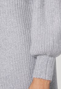 Vaaleanharmaa neulepusero, jossa on pystysuoraa ribbausta, pullistuneita hihoja ja pehmeä rakenne. Lähikuva hiuksen yksityiskohdasta.