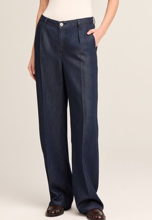 Woman wearing dark blue wide-leg denim pants with front pleats, button closure, side pockets, and brown leather shoes, hands in pockets.