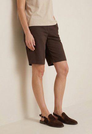 Person iført mørkebrune knælange shorts, beige kortærmet top og mørkebrune fløjlsbage sko, stående mod en lys væg.