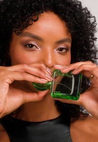 Grønn glass parfymeflaske holdt av en person, med kvadratisk form, glatt overflate og preget tekst, delvis fylt med væske.