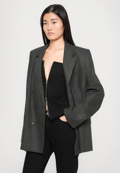 Blazer grigio doppiopetto con righe bianche, vestibilità oversize, spalle strutturate, grandi bottoni, abbinato a un top nero con zip e pantaloni aderenti.