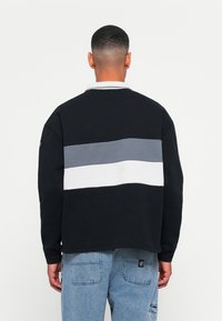 Man in zwarte trui met grijs en witte horizontale strepen op de achterkant, gecombineerd met lichtblauwe jeans, staande met zijn rug naar de camera op een effen achtergrond.