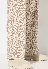 Vidjebyxor i krämfärg med ett brunt lövmönster, gjorda av texturerat tyg, kombinerade med beige klumpiga skor.