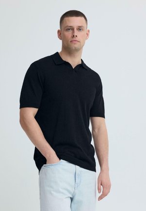 Ung mand med lys hud og kort brunt hår iført sort kortærmet poloshirt og lyseblå jeans, stående mod en ensfarvet baggrund.