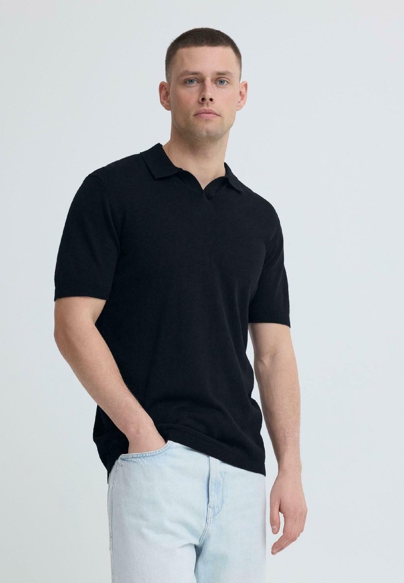 Νέος άνδρας με ανοιχτόχρωμο δέρμα και κοντά καστανά μαλλιά φοράει μαύρο polo με κοντά μανίκια και ανοιχτό μπλε τζιν παντελόνι, στέκεται μπροστά σε ένα απλό φόντο.