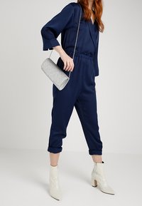 Giocattolo blu navy, tuta a maniche lunghe con vita elastica, abbinata a una borsa a mano glitterata argento e stivaletti bianchi con tacco.