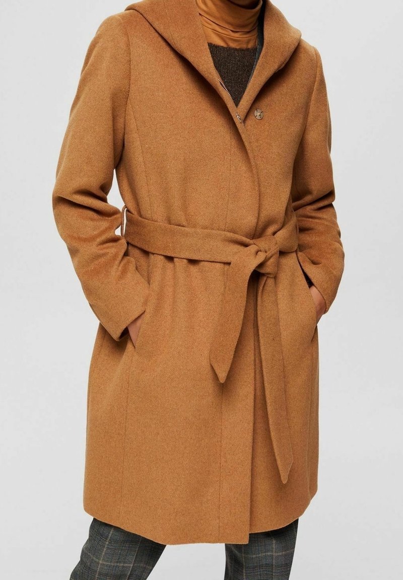 Camel-kleurige wollen jas met een gestrikt ceintuur, sjawlboord en handen in de zakken, gedragen over een bruine coltrui en geruite broek.