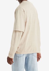 Osoba w beżowej, warstwowej koszuli z długim i krótkim rękawem oraz jasnoniebieskich dżinsach, widoczna od tyłu.