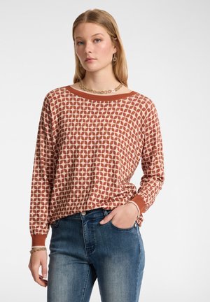 Blusa de manga larga en rosa claro con un patrón de hojas marrón repetido. Presenta un escote barco y puños acanalados, combinada con jeans azules.